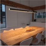 Contemporary black hanging linear light with remote providing warm illumination over a long light wood dining table.